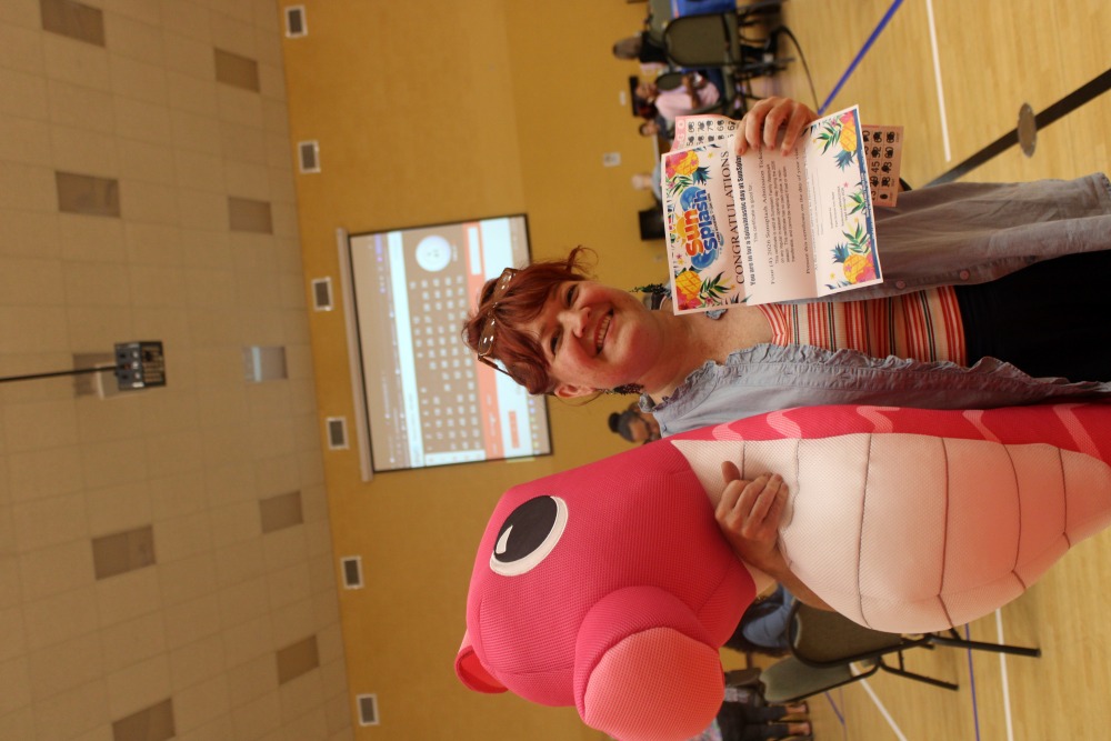 Smiling woman at South Cape Community Center holds a large pink inflatable seahorse and a family bingo prize certificate in front of the projected bingo screen